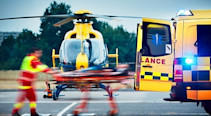 A paramedic pushing a stretcher into an ambulance with an air-ambulance helicopter in the background.