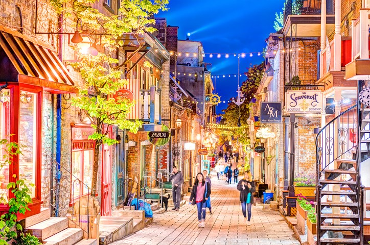 Several people walk down the narrow Rue du Petit-Champlain at dusk, with the street’s various shopfronts joined and illuminated by strings of lightbulbs.