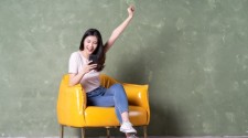 A woman sat on a sofa looking excited while holding a mobile phone.
