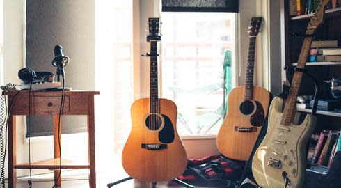 Two acoustic guitars and one electric guitar stand upright between a half-full bookshelf and a small wooden desk on which a professional microphone and headphones are placed.