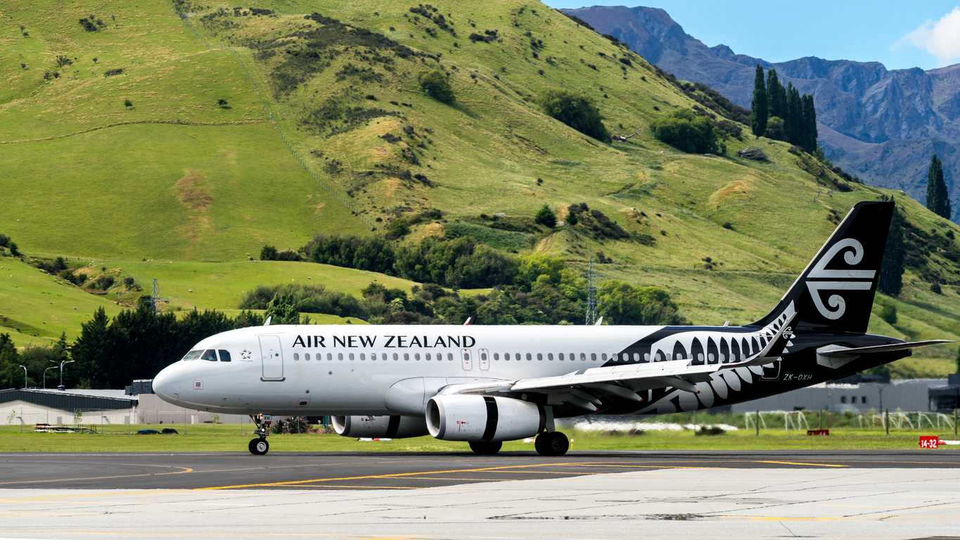 Pesawat Air New Zealand tengah bersiap lepas landas dengan latar belakang bebukitan yang rindang