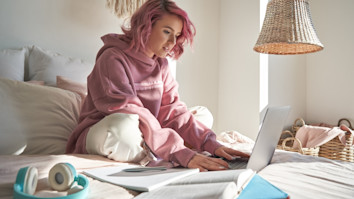 Student with pink hair wearing hoodie using laptop while sitting in bed 