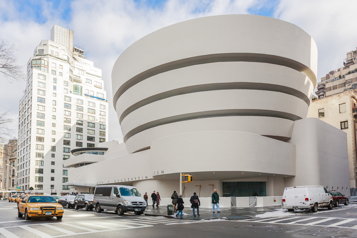 The striking exterior of the Solomon R. Guggenheim Museum in New York City, with its four cream concentric circle roofing elements standing beside pedestrians, vehicles and other tall buildings