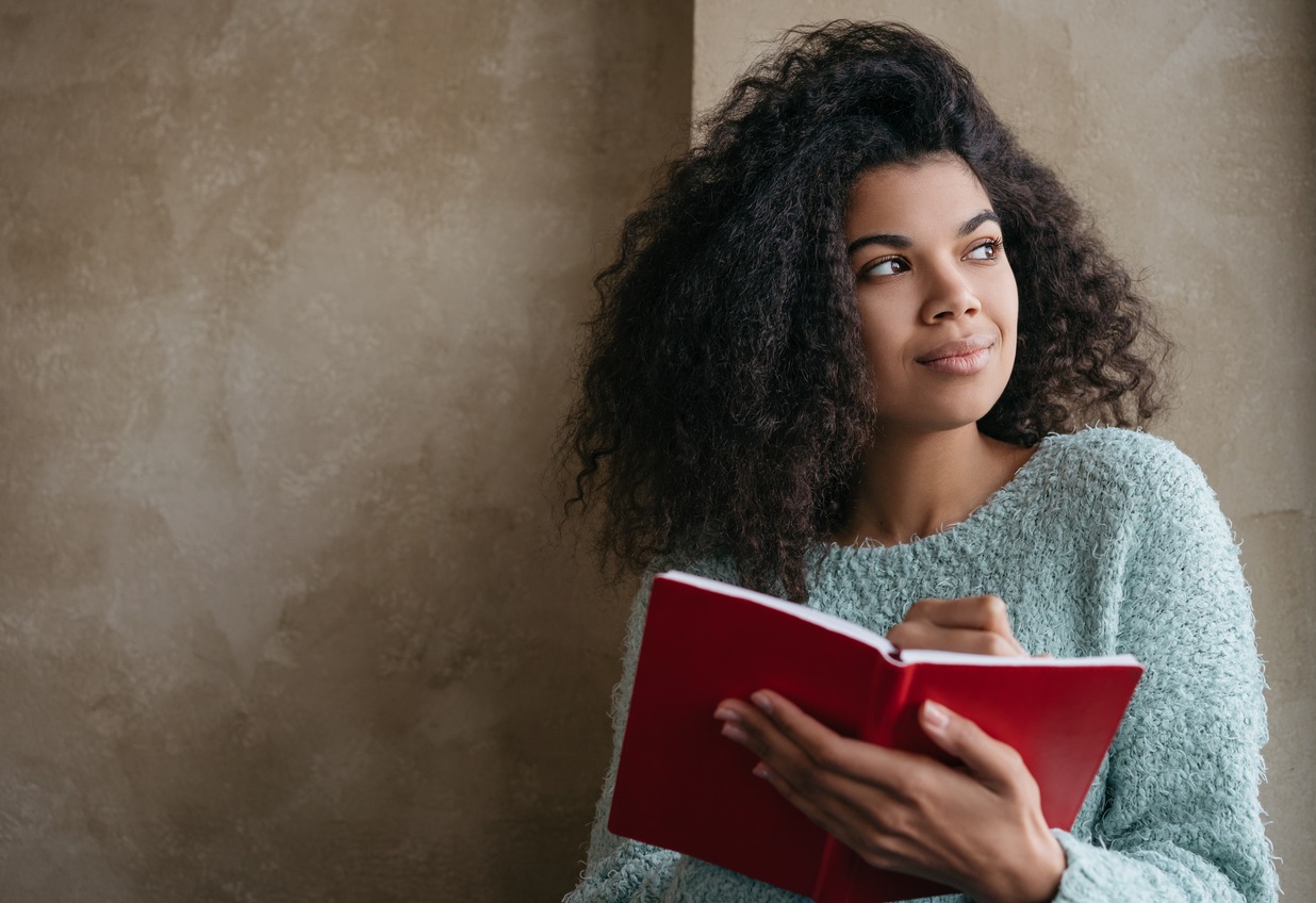 Young woman in blue jumper looks to her left while holding a red notebook. She smiles slightly as if coming to a realisation.