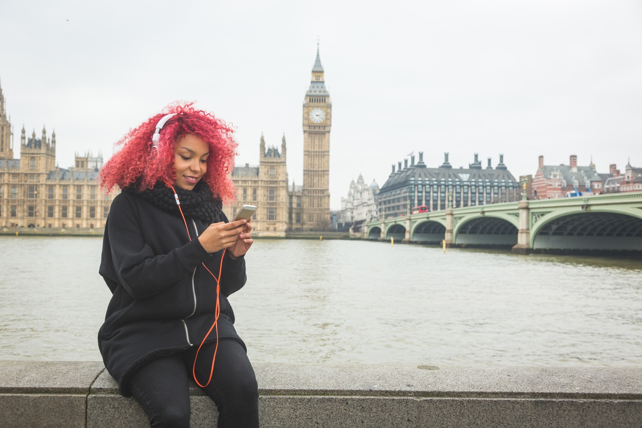 Young woman smiles while sat on a wall over the south bank of the Thames. She is looking at her phone and wearing headphones with Westminster behind her