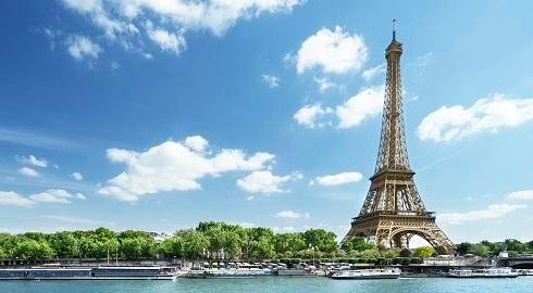 The Eiffel Tower stands tall over green trees and small cruiser boats on the Seine river beneath blue skies in Paris