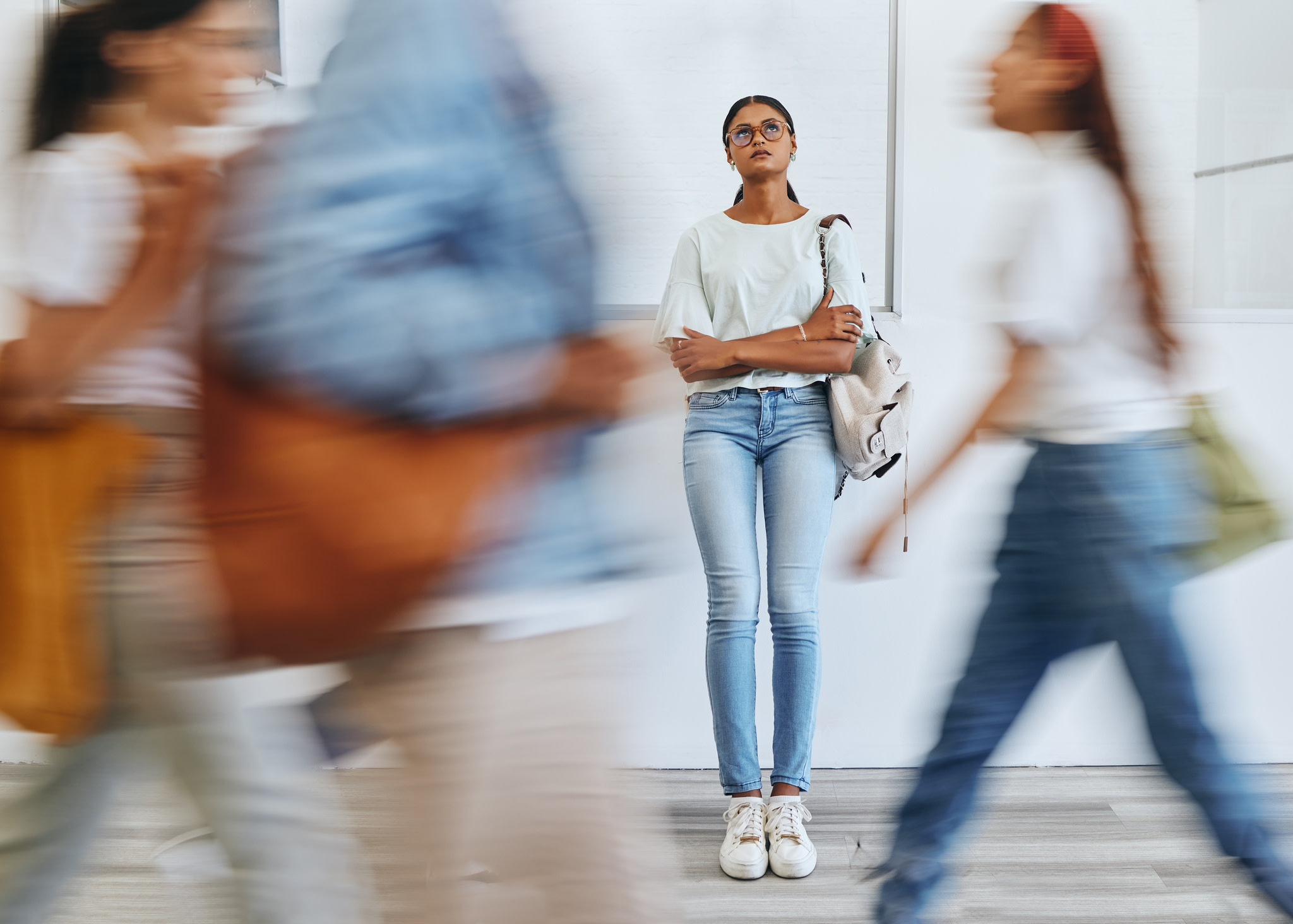 Young woman with a bag over her shoulder looks upwards wistfully from the back of a corridor through which a blur of three other people are walking