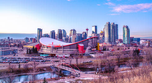 The Calgary skyline at sunset, with the Olympic Saddledome in the foreground and several skyscrapers in the distance behind it.