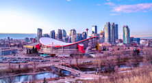The Calgary skyline at sunset, with the Olympic Saddledome in the foreground and several skyscrapers in the distance behind it.