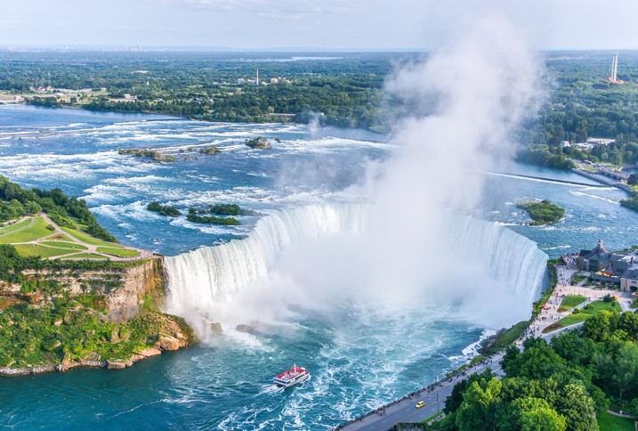 A boat tour full of people sails towards the fast-rushing Horseshoe Falls on the Niagara River. Tall green trees fill the shoreline and extend a long way inland.