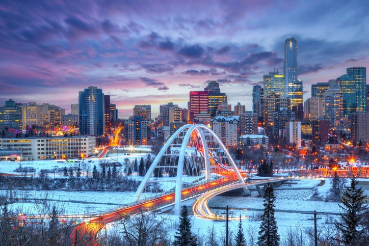 Walterdale Bridge stands over the frozen Saskatchewan River in front of the many tall buildings of the Edmonton skyline, all lit up at dusk.