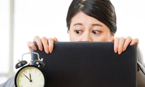 Foto pelajar yang menyembunyikan wajahnya di balik laptop sambil melirik ke arah jam