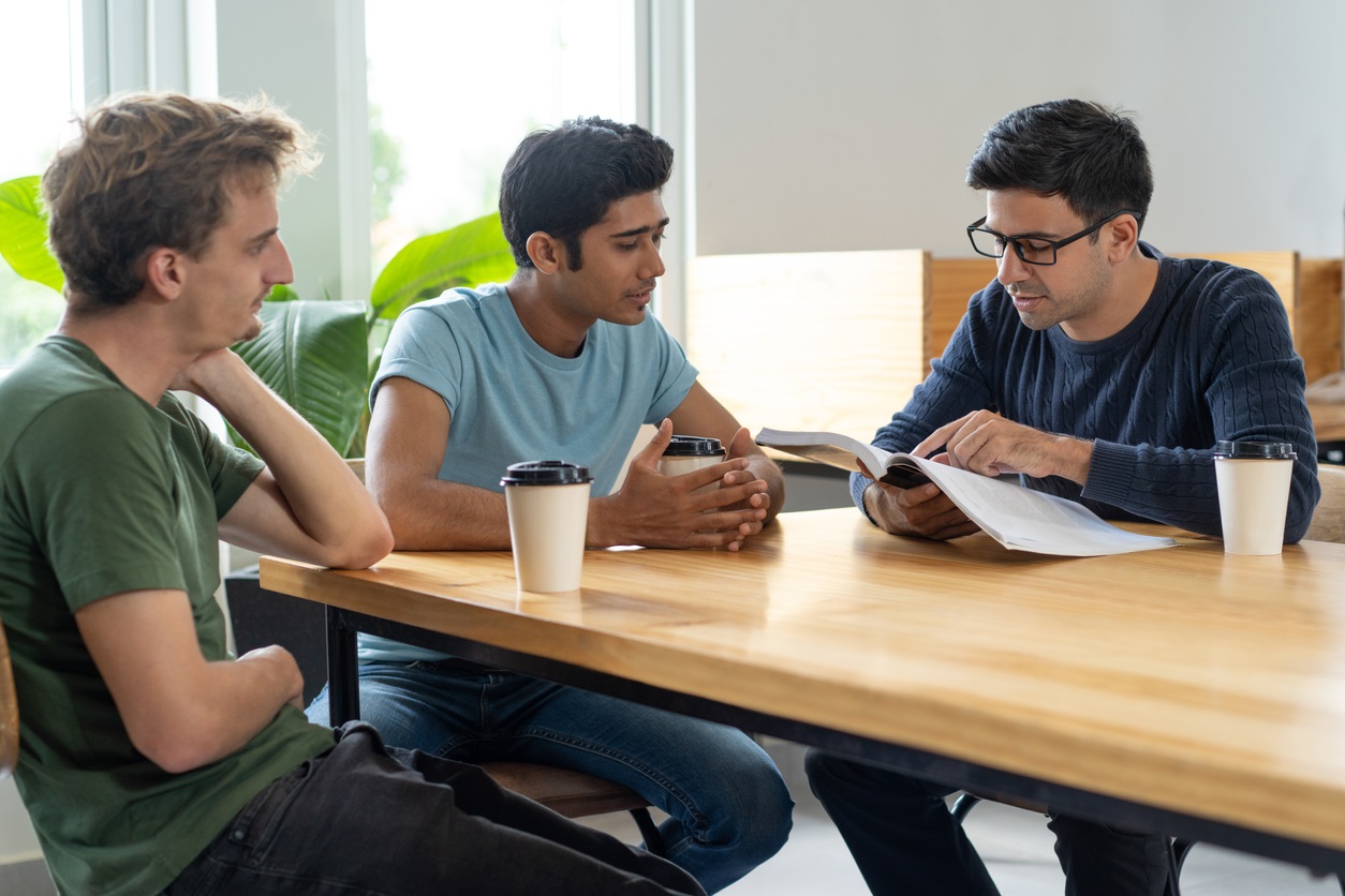 Three male students sit around a wooden table as one points out something in a book and the others look towards him. All three have disposable coffee cups.