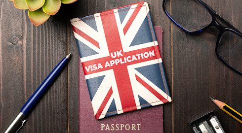 A small folder with a Union Flag design bearing the words 'UK visa application' rests on top of a passport and a wooden table. Beside it are a pen, pencil, pencil sharpener and eyeglasses