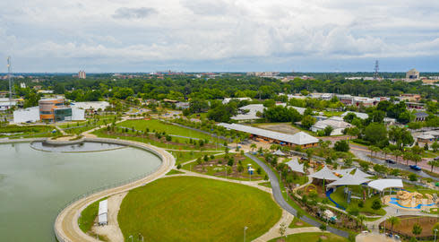 Aerial view of Depot Park in Gainesville, with a man-made lake beside green lawns, and several seating and play areas. Tall green trees stretch into the distance beneath a cloudy blue sky.