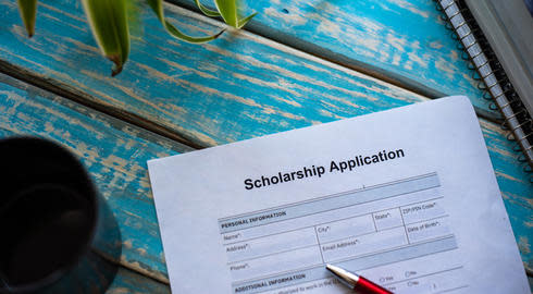 Scholarship application form on blue wooden table. There's the tip of a red pen on the form. Green leaves in the top left hand corner. Black notebook in top right corner. 