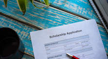 Scholarship application form on blue wooden table. There's the tip of a red pen on the form. Green leaves in the top left hand corner. Black notebook in top right corner. 