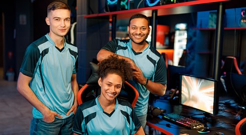 Three players for an esports team smile as two stand and one sits beside a gaming computer
