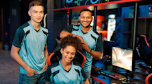 Three players for an esports team smile as two stand and one sits beside a gaming computer