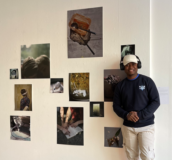 Emmanuel Sandy smiles while wearing headphones as he stands by a white wall covered in several artistic photos