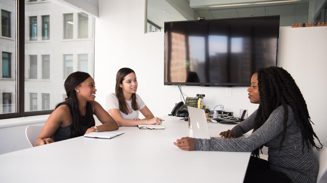 Three women talking at conference table with laptop and notebooks