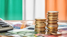 Piles of Euro coins and banknotes rest next to a calculator in front of the flag of Ireland