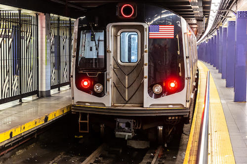 New York subway train with two lit red lights and a USA flag on its front end, stationary between two platforms