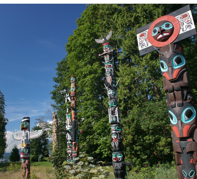  Colourful Totem Poles in Stanley Park, BC Vancouver stand in the foreground. Behind the poles are tall green trees with blue skies