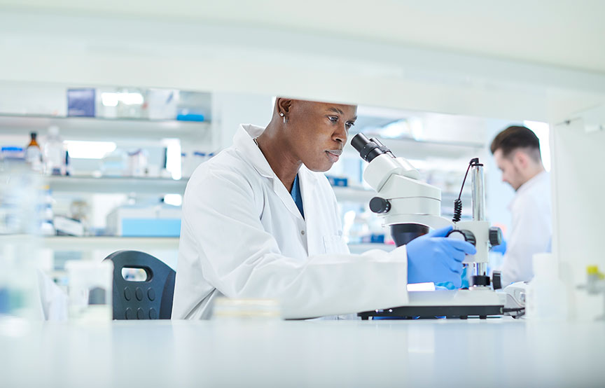 woman wearing a white lab coat and blue gloves in a lab looking into microscope