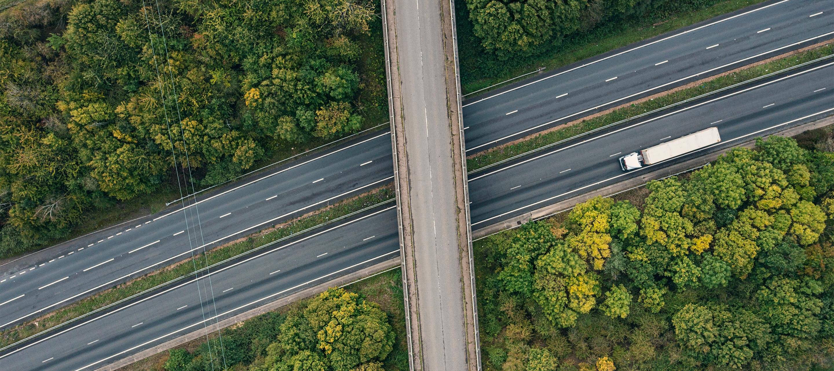 image shows an aerial view of a mostly empty highway with green trees surrounding the road, one white truck is seen driving through