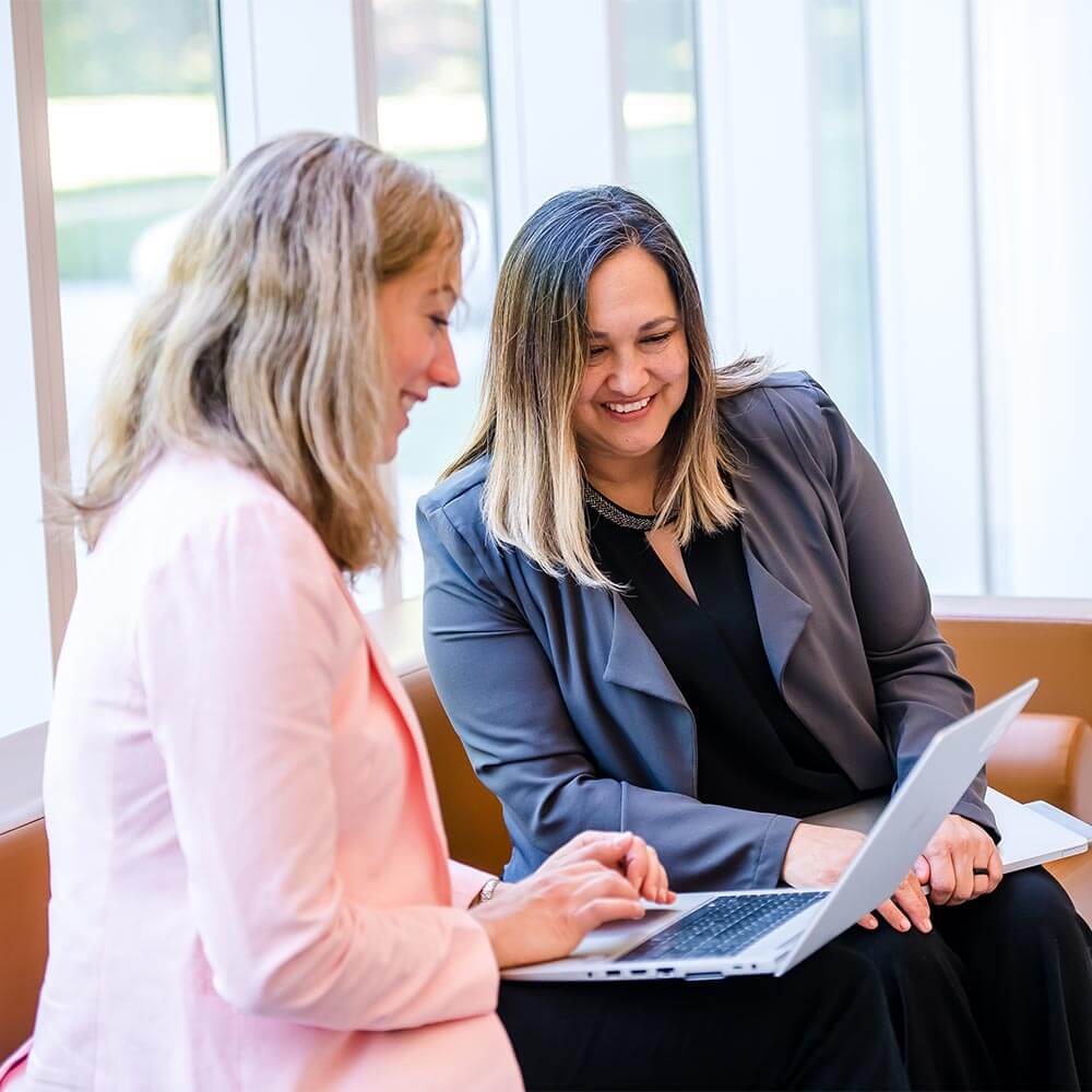 two women sitting together and smiling at a laptop screen