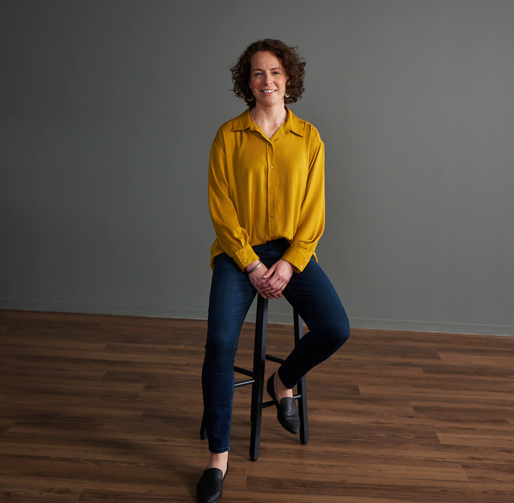 woman in yellow shirt by a grey wall, sitting on stool 
