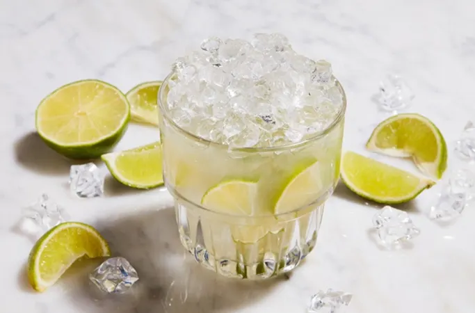 A caipirinha cocktail on a marble background surrounded by lime wedges and ice