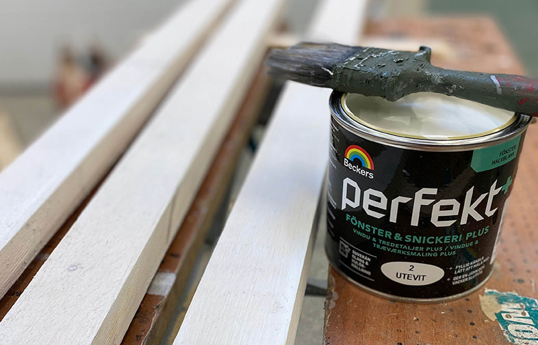 Svart färgburk med vit färg och pensel på arbetsbänk, omgiven av vita träplankor.