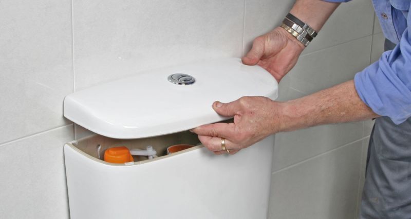 Händer lyfter vitt toalettcisterntak i kaklat badrum, synliga orange delar inuti.