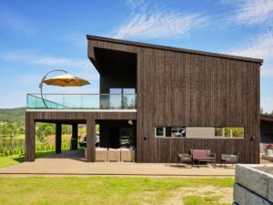 Modern mörkbrun trävilla med glasräcke, gul parasoll på uteplats, grön gräsmatta och vinodling.