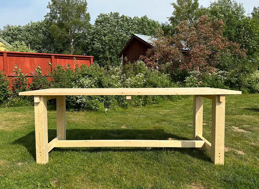 Ljust träarbetsbord på grön gräsmatta framför rött staket och röd stuga bland buskar