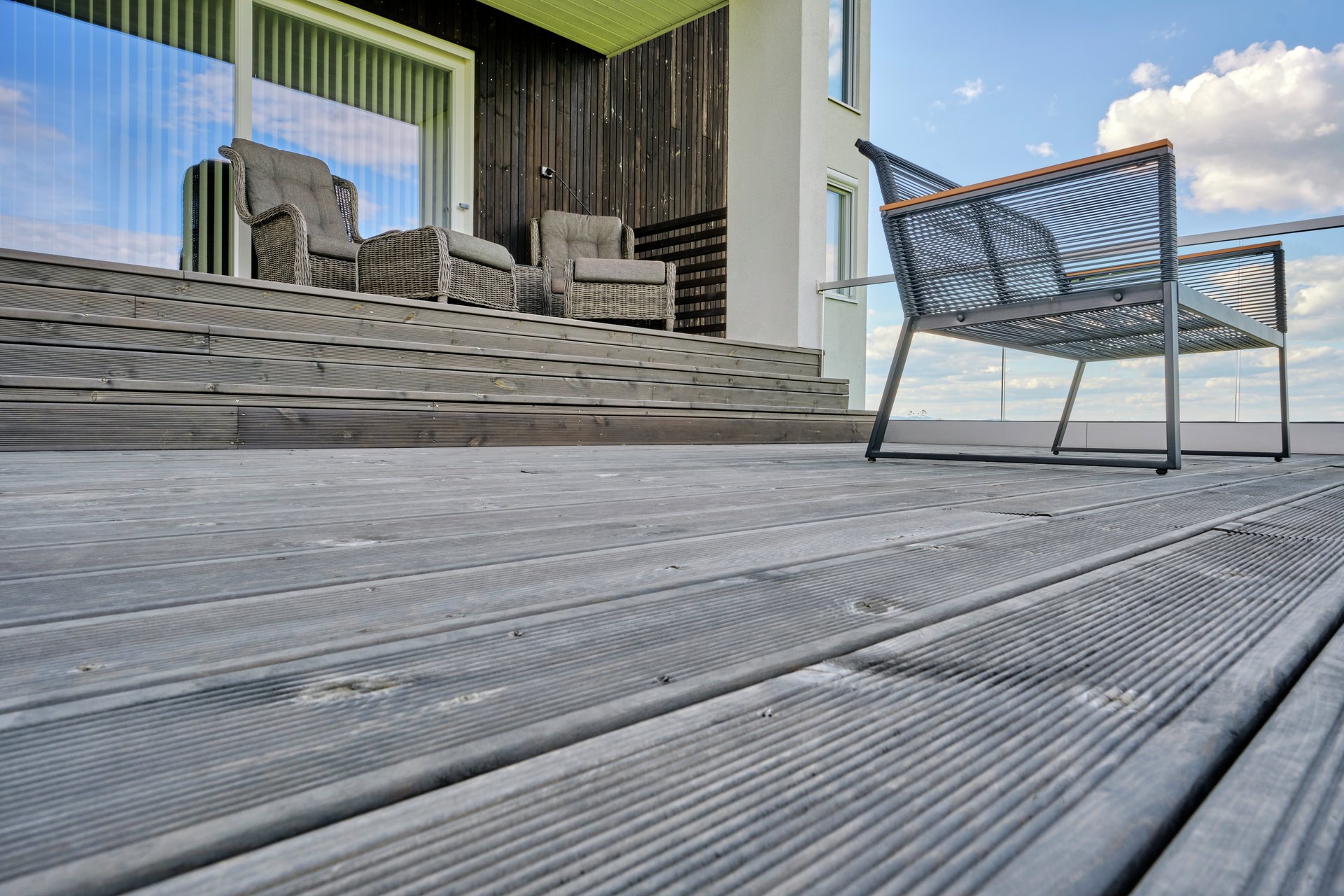 Terrass med grå trall, svart metallstol i förgrunden och två grå rottingfåtöljer