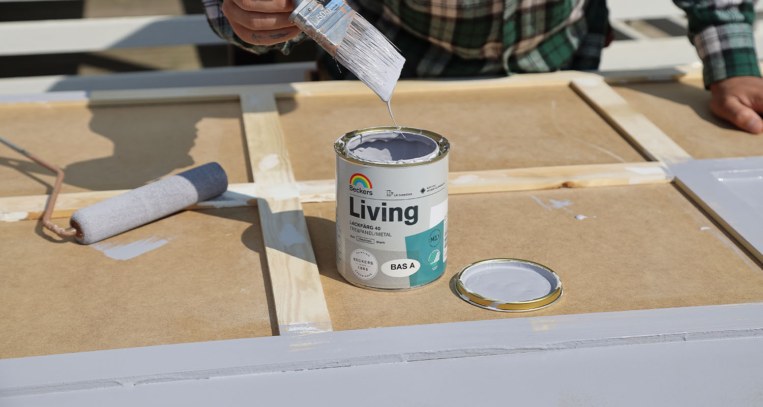Hand med pensel droppar grå Beckers Living-färg på träram, roller bredvid.