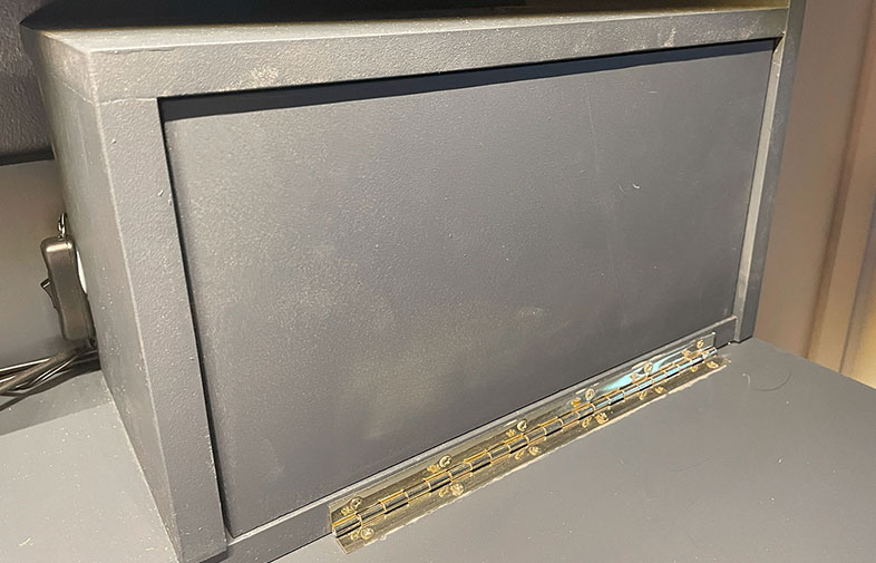 Mörkgrå rektangulär låda med guldigt gångjärn längst ner, på grå bordsyta.