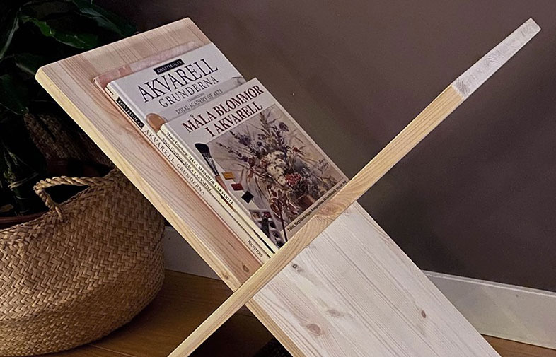 Ljust trästativ med två akvarellböcker på bord, flätad korg med grön växt bakom