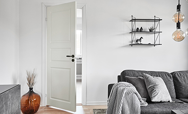 Vitt vardagsrum, öppen vit dörr, grå soffa med pläd, vägghylla med figurer, hängande lampor och vas.