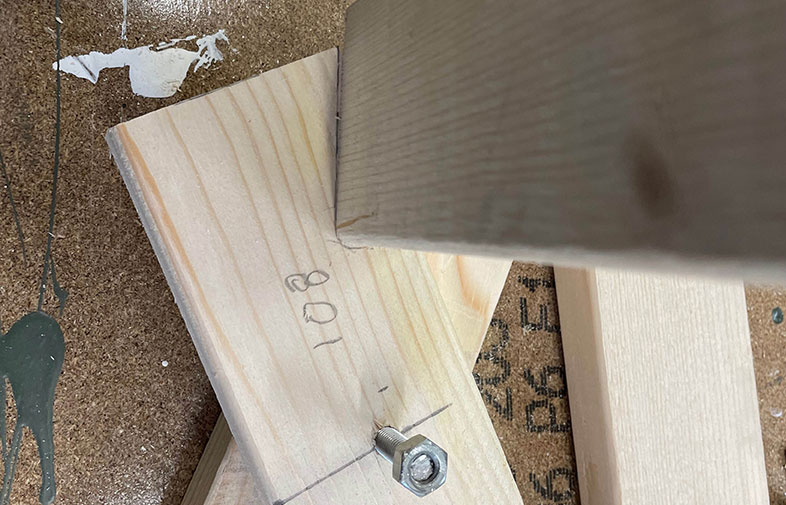 Två ljusa träplankor i hörnfog på mörkt arbetsbord med färgstänk, märkning “101” och mutter.