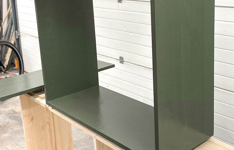 Mörkgrönt U-format hyllplan på träbockar i verkstad med vit liggande brädvägg.