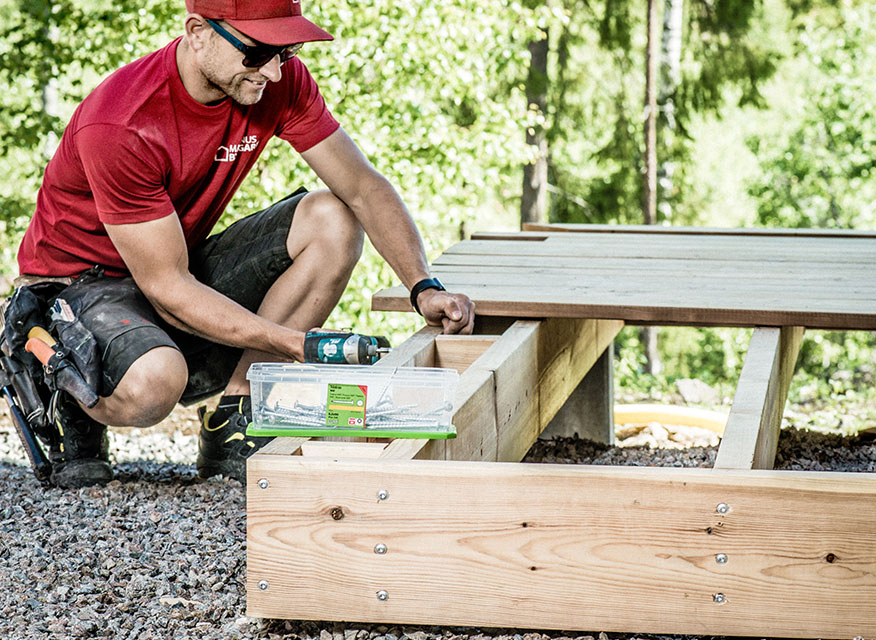 Person i röd tröja monterar trallram av trä på grus med borr, grön skog i bakgrunden.