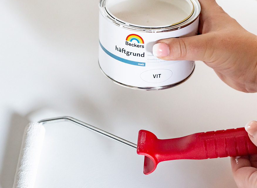 Hand håller öppen burk vit häftgrund från Beckers och röd roller på vit bakgrund.