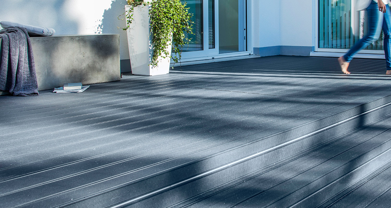 Mörkgrå terrass med komposittrall i solljus, skuggor, betongbänk, kruka och barfota ben