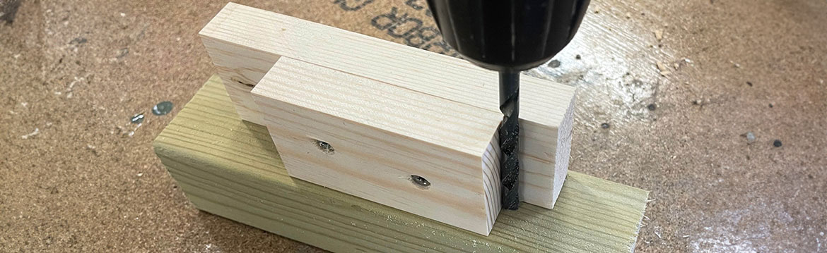 Närbild på svart borr som borrar i två ljusa träbitar monterade på en grön träkloss.