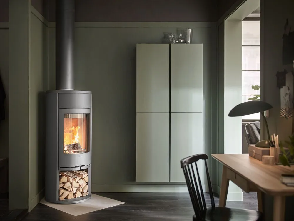 Grönt rum med svart rund vedkamin, vedförvaring, ljusgröna skåp och arbetsbord.