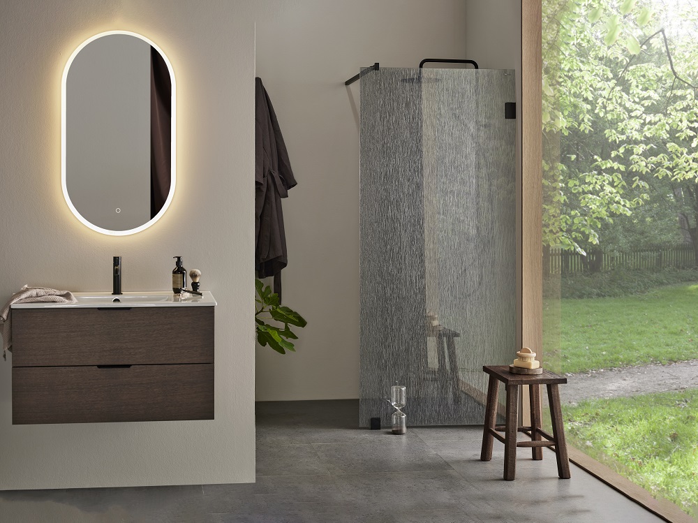 Modernt badrum med mörk träkommod, upplyst oval spegel, dusch bakom mönstrat glas och grön utsikt.